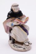 A Bow figure of a monk wearing a black and pink robe, seated on a stool reading a large Bible, a