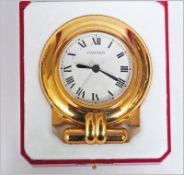 Cartier gilt metal travelling alarm clock, the circular white dial with Roman numerals, on folding