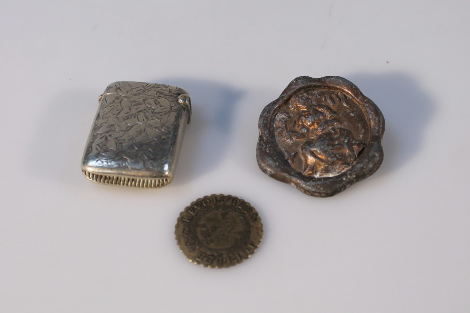 A silver plated small vesta, a brass gaming token, and a badge.