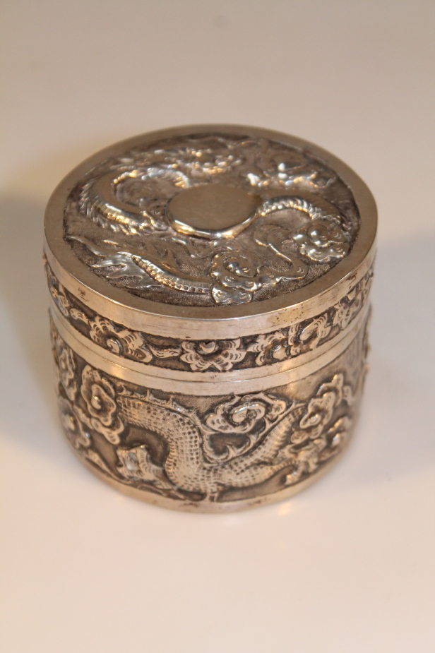 A Chinese white metal circular trinket box, repoussé decorated with dragons, stylized clouds,