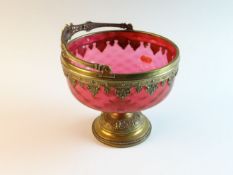 An early 20th Century cranberry glass and brass mounted bowl with hinged handle on stepped