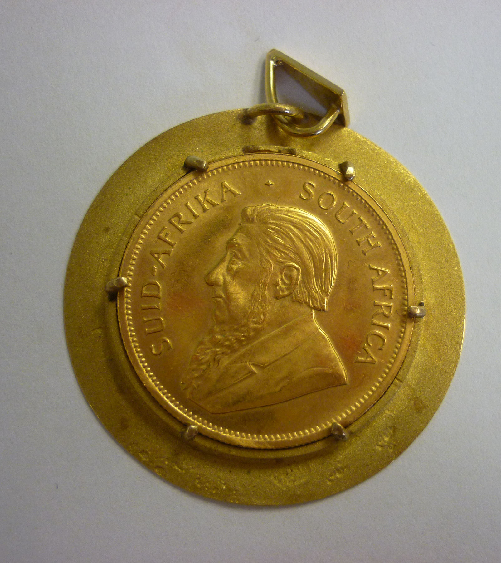 A 1977 krugerrand, set in a textured yellow metal frame with a pendant loop