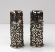 A pair of white metal pepperettes, with a pierced domed cover, the cylindrical body embossed with