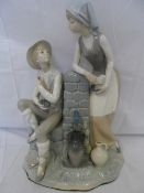 A LARGE LLADRO STYLE FIGURE OF A BOY AND GIRL LEANING AGAINST A WATER WELL, APPROX. 35 X27 CM