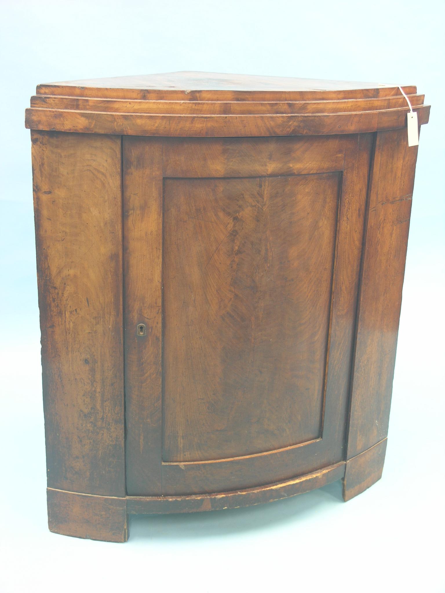 An early 19th century mahogany corner cupboard, enclosed by single bow-front door with flame veneer,