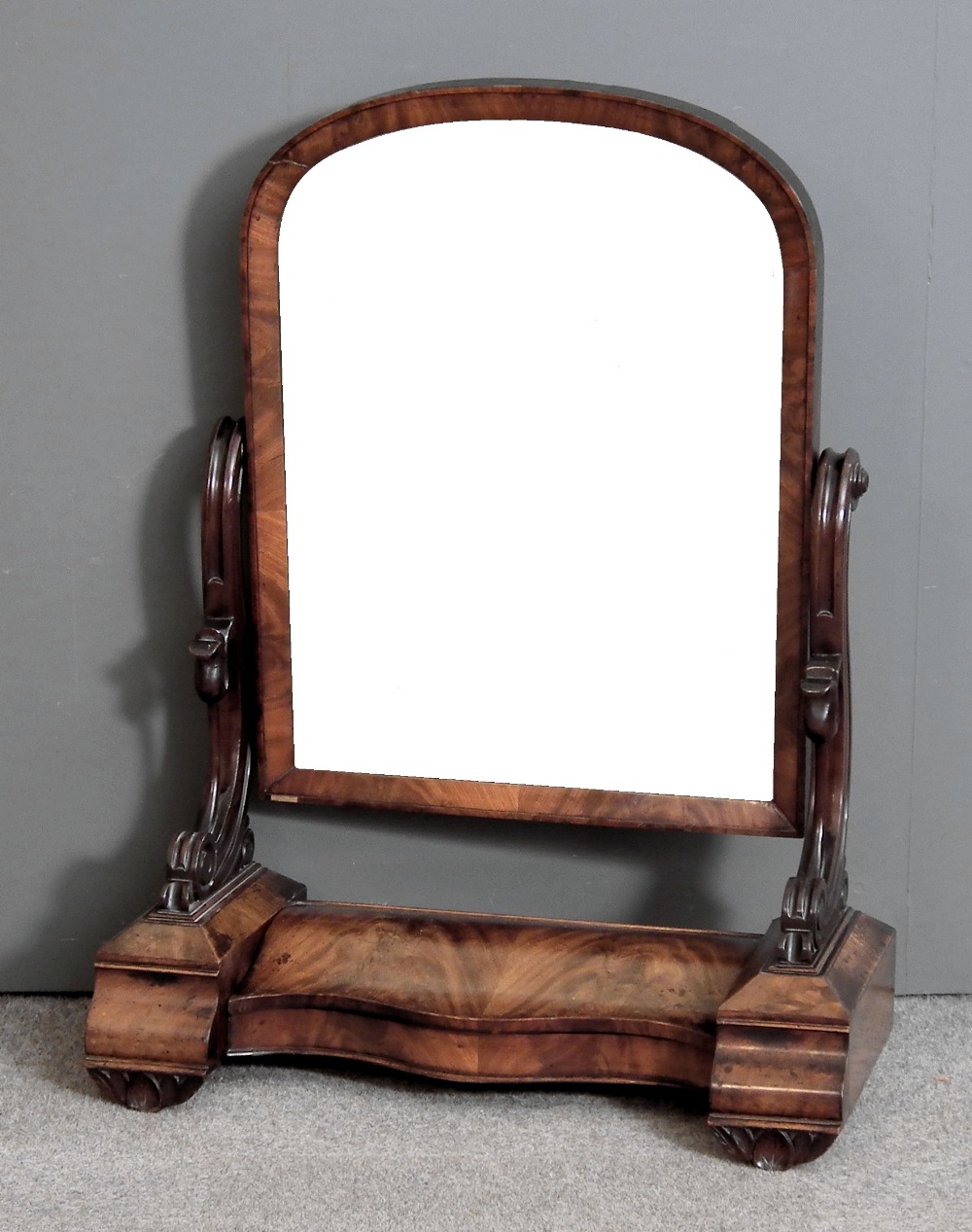 An early Victorian figured mahogany rectangular toilet mirror with arched top, on shaped supports,