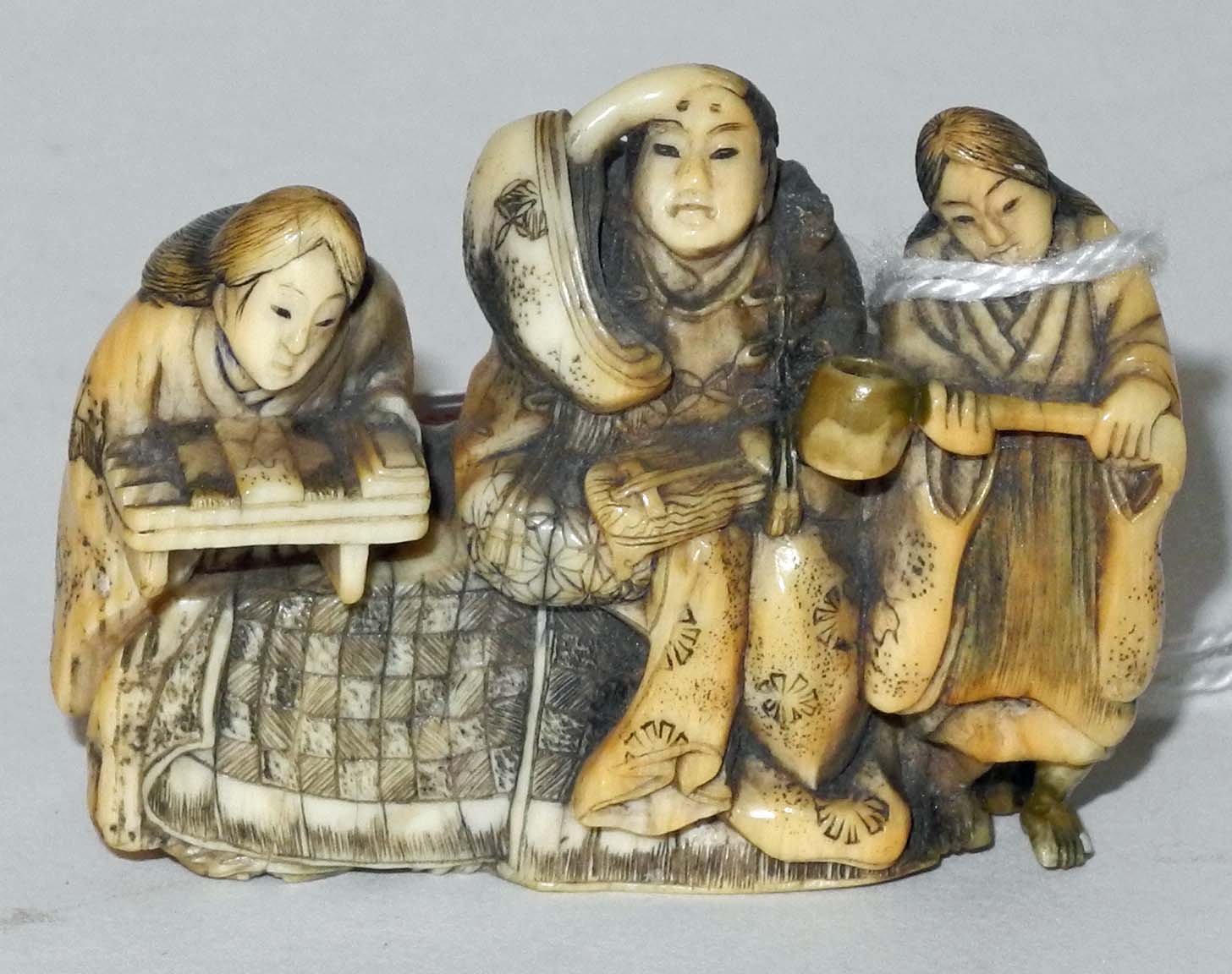 A Japanese ivory netsuke of three figures, depicting a domestic scene, two seated, with rope