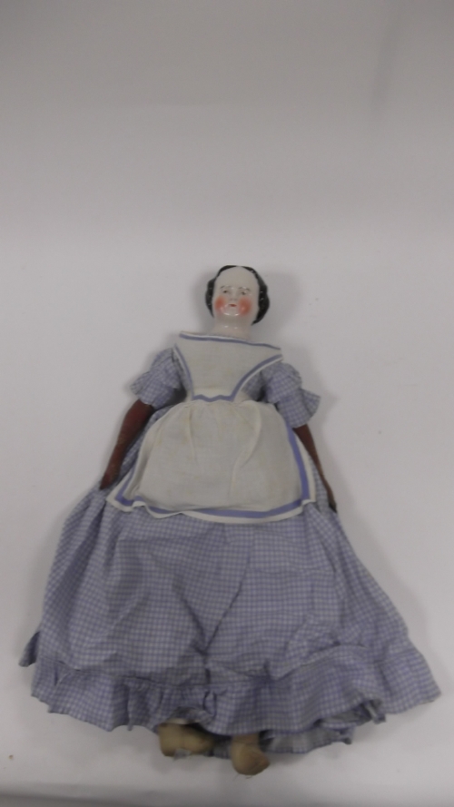 A porcelain headed doll with brown eyes and leather body, wearing a blue check dress with apron,