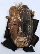 A carved wooden African tribal warrior mask bearing bird feathers to the forehead with a cloth
