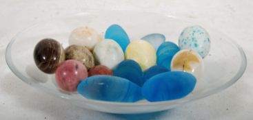 A collection of 16 glass and stone eggs together in a glass bowl measuring 36cm diameter.