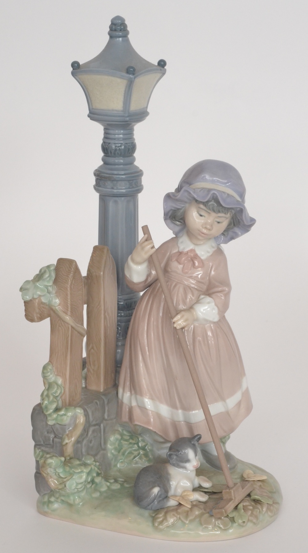 A coloured Lladro figure `The Clean Up` modelled as a young girl sweeping beside a lamp post,