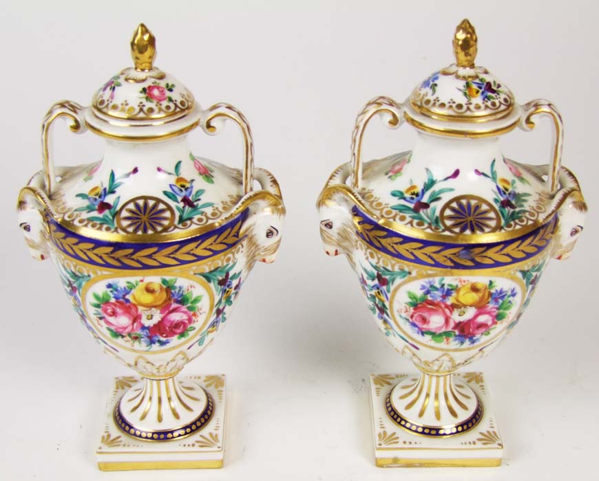 A pair of porcelain twin handled urns and covers, with goat masks, and panels of flowers, on