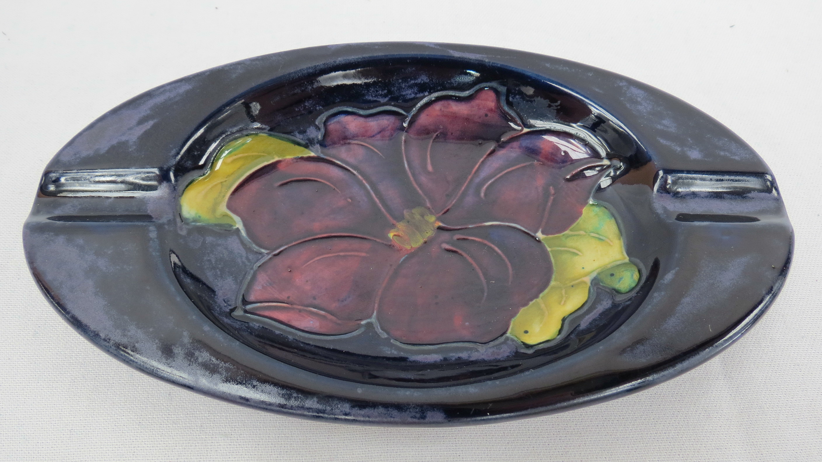 A Moorcroft oval ashtray decorated with a clematis flower on a dark blue ground, measuring 16cm