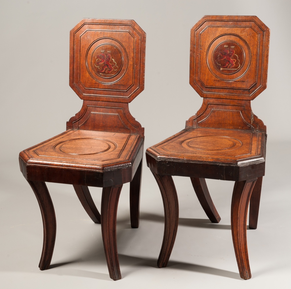 A pair of Regency mahogany hall chairs, each with carved panel back centred with painted crest of