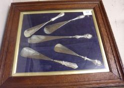 An Oak Surround and Glazed Frame holding five assorted steel bladed and silver handled Shoe Horns,
