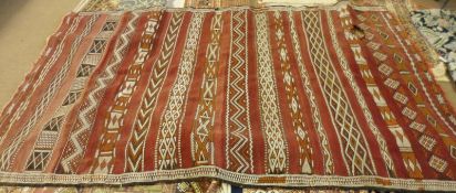 A 20th Century Kelim Carpet, decorated with panels of geometric designs on a red field, (holed and