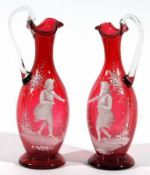 Two Cranberry Ewers each with clear looped handles and painted in the manner of Mary Gregory with