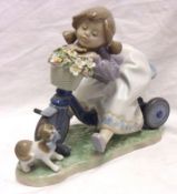 A Lladro Model of a young girl on a trike with a basket of flowers and a kitten below, 7 ½” long