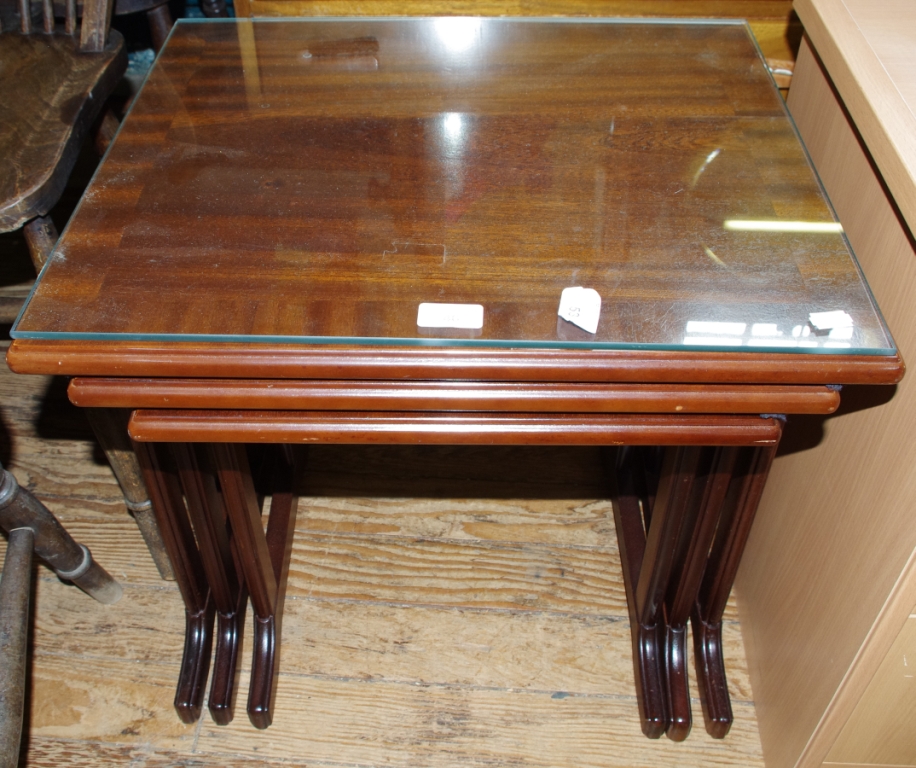 An oblong shaped mahogany nest of three tables with trestle supports and glass tops