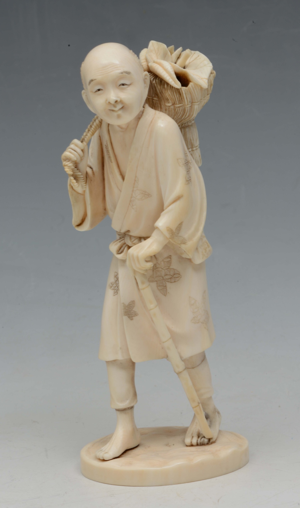 A JAPANESE IVORY OKIMONO in the form of a farmer walking with a bamboo cane and carrying a sheath of