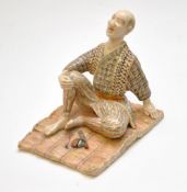 A Japanese Satsuma figure of a man wearing striped trousers sat before a pipe and bag on a rug, gilt