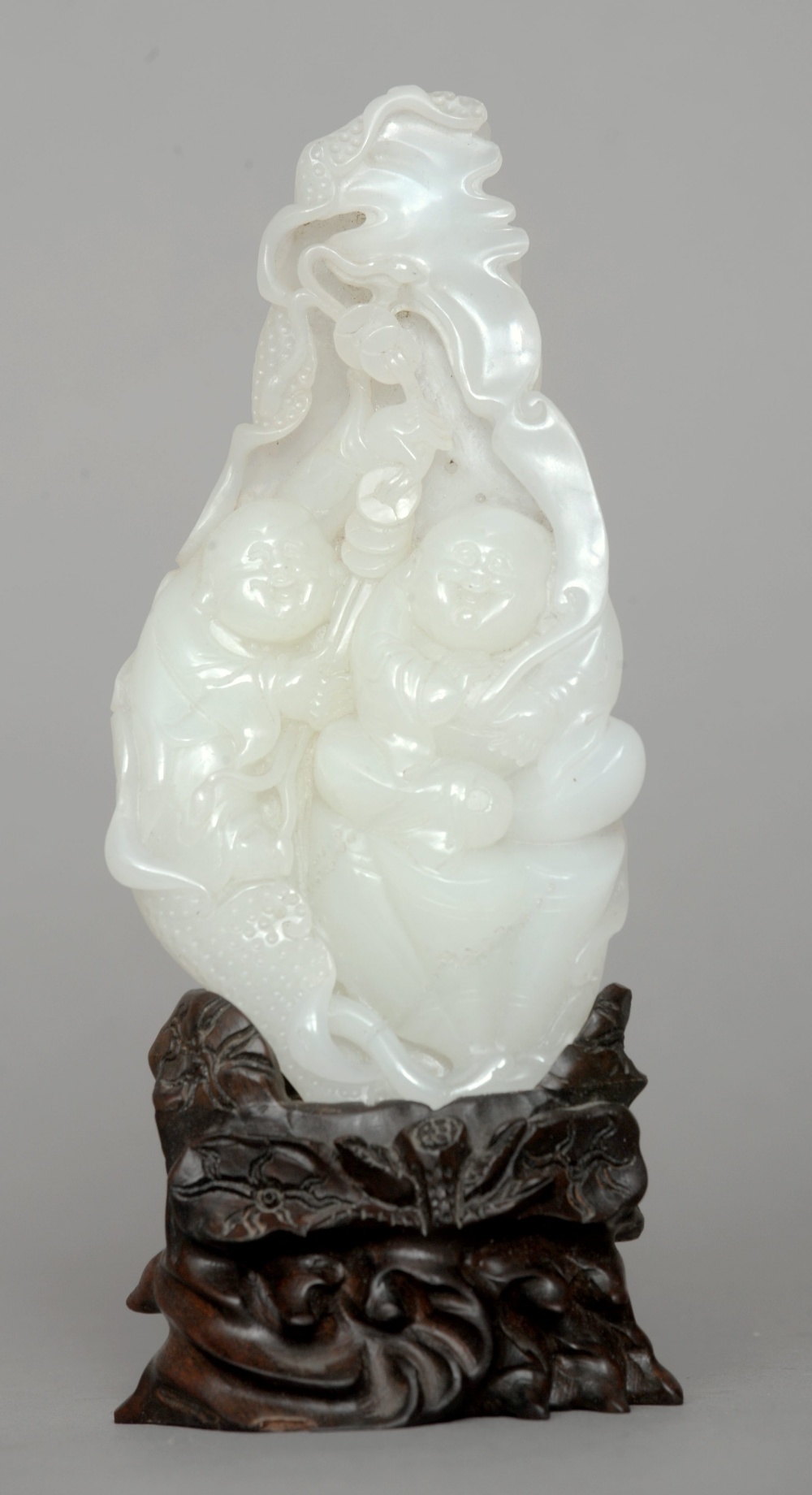 A Chinese white jade group Carved as two laughing boys with lucky charms, seated within a foliate
