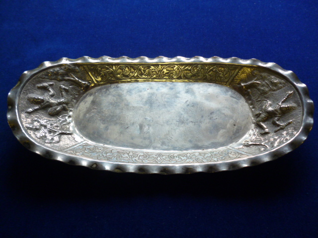 A Far Eastern white metal dish, oval shaped with wavy rim and raised design of figures and trees,