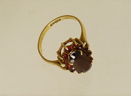 A 9ct gold garnet set dress ring, the ring set with a central oval faceted garnet with a surround of