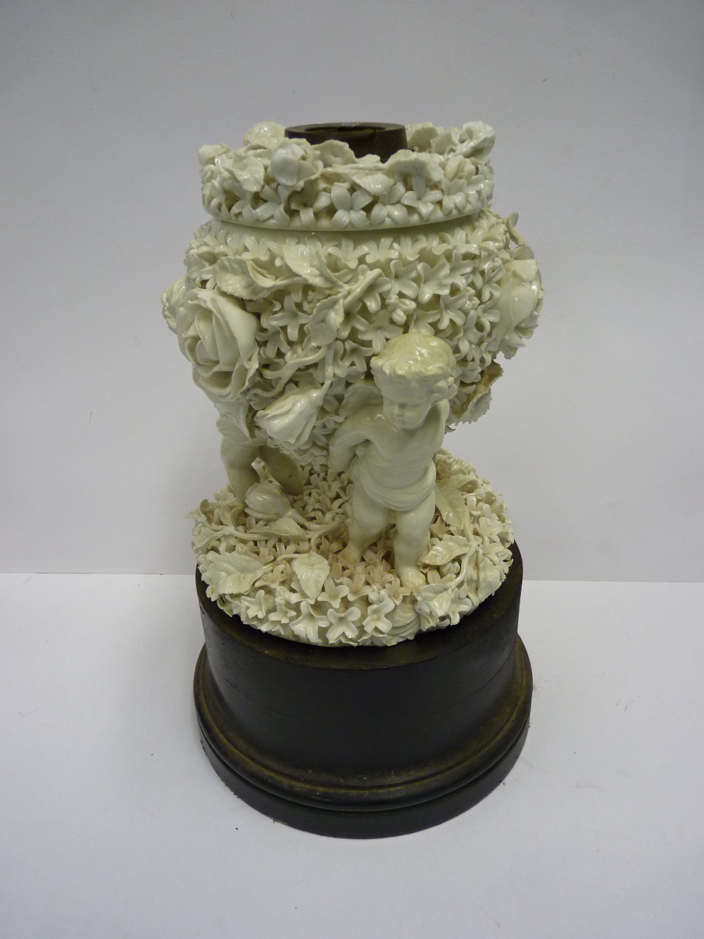 A glazed white ceramic oil lamp base, modelled as three cherubs holding up a flower encrusted oil