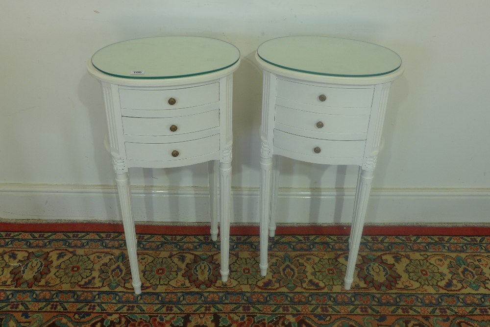 A pair of oval white painted three drawer glass topped bedside chests on fluted legs 72cm high x