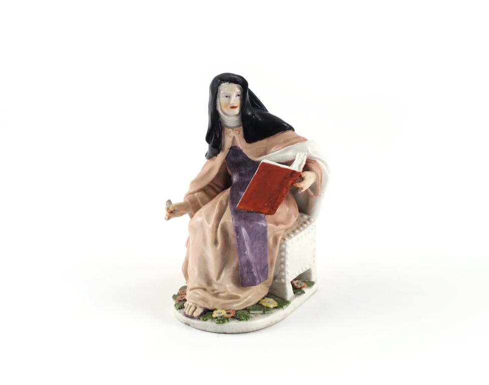 A Chelsea figure of a nun c.1755, seated on a studded chest and reading from a large book