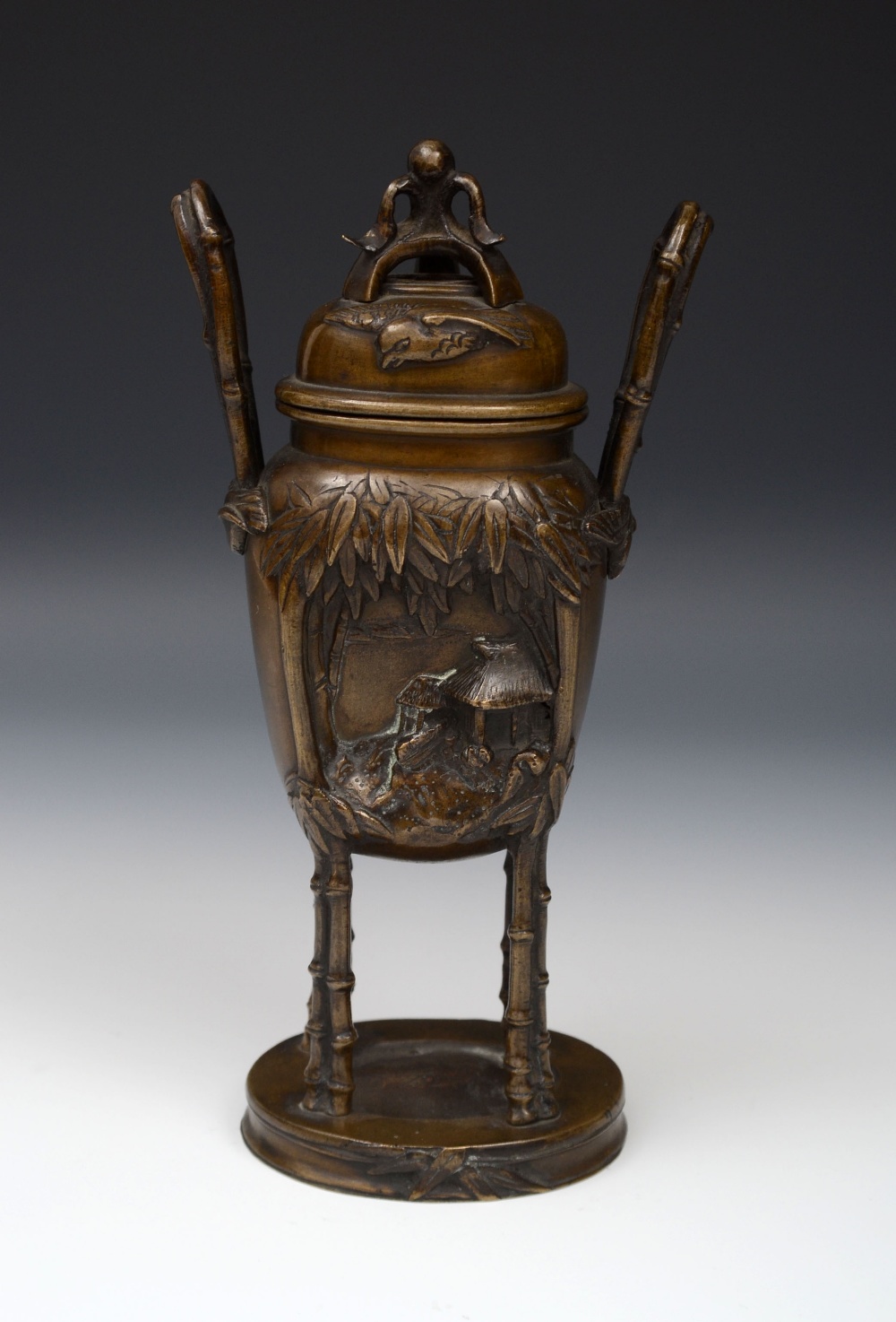 A Japanese patinated bronze two handled censer, cast with figures and houses, raised on stylised