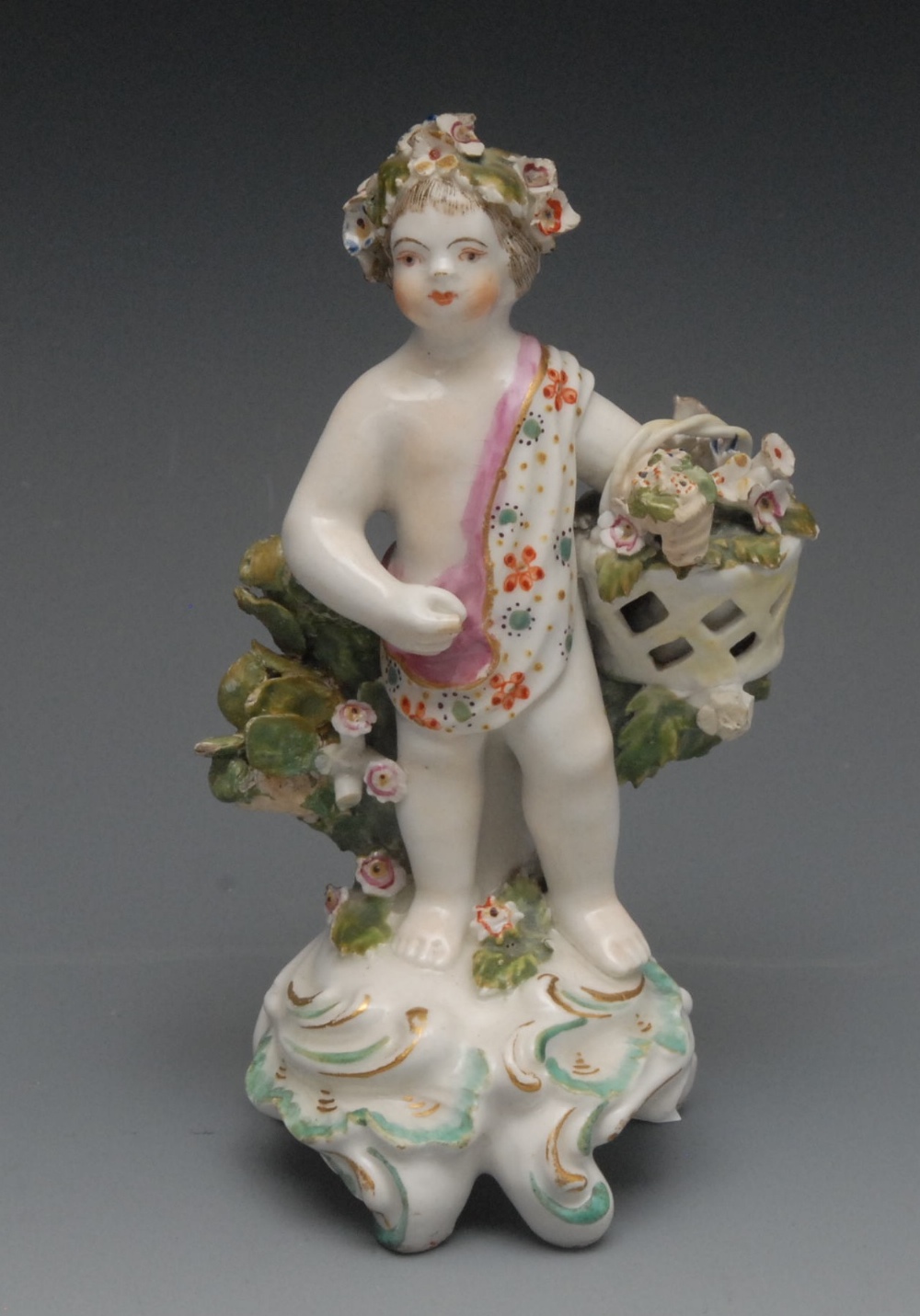 A Bow figure, of a putto, standing scantily draped wearing a floral chaplet, holding a basket,