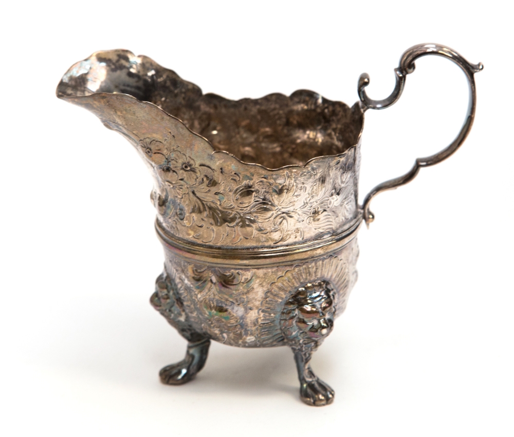 A George III Irish silver milk jug of Grecian helmet form on three lions head and paw feet, embossed