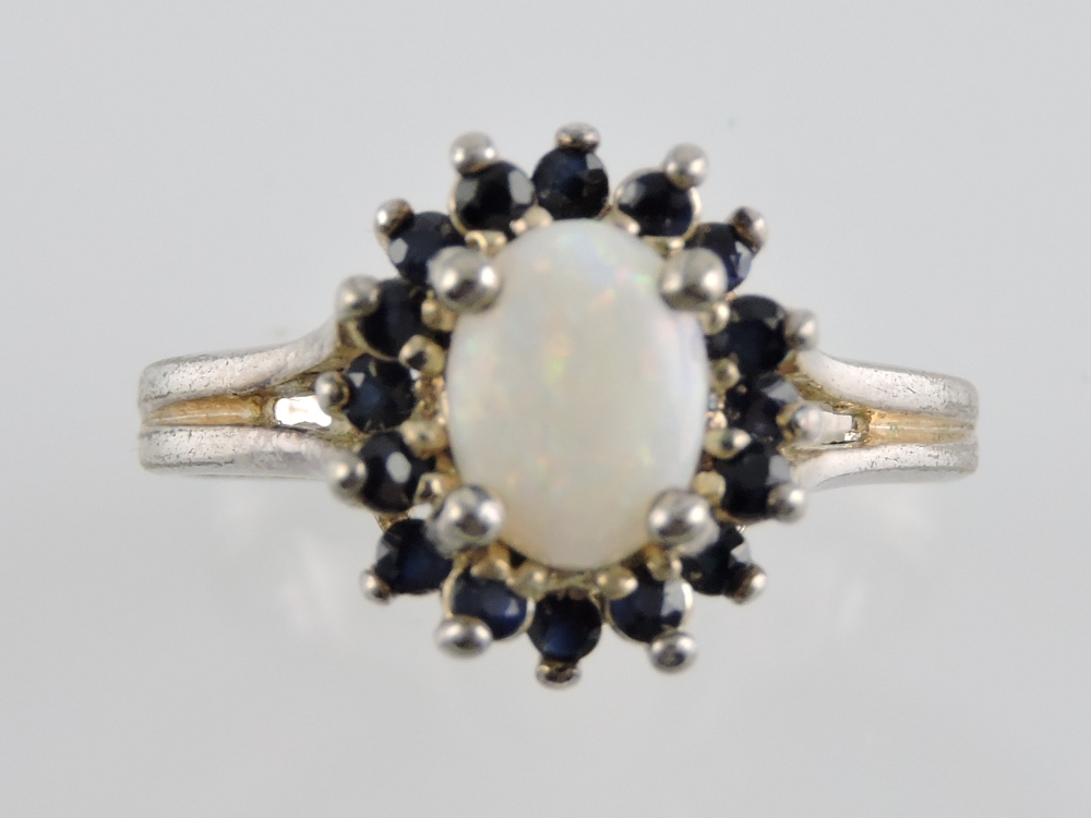 An opal and sapphire dress ring, the oval central stone within a band of claw set stones, on a white