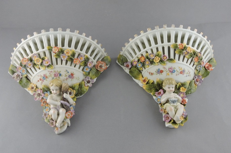 A pair of continental porcelain floral encrusted bow front wall brackets moulded with seated putti.