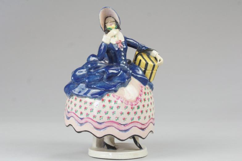 An art pottery crinoline figure by Kathy Evershed, in a standing pose holding a flower and a hat
