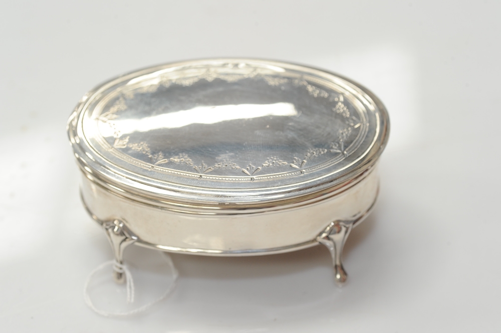 A silver trinket box of oval form and on four moulded supports, the hinged lid with engraved