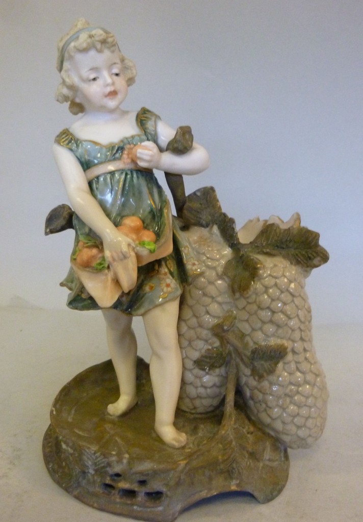 A late 19thC Ernst Wahliss porcelain spill vase, fashioned as a young girl, standing beside a grape
