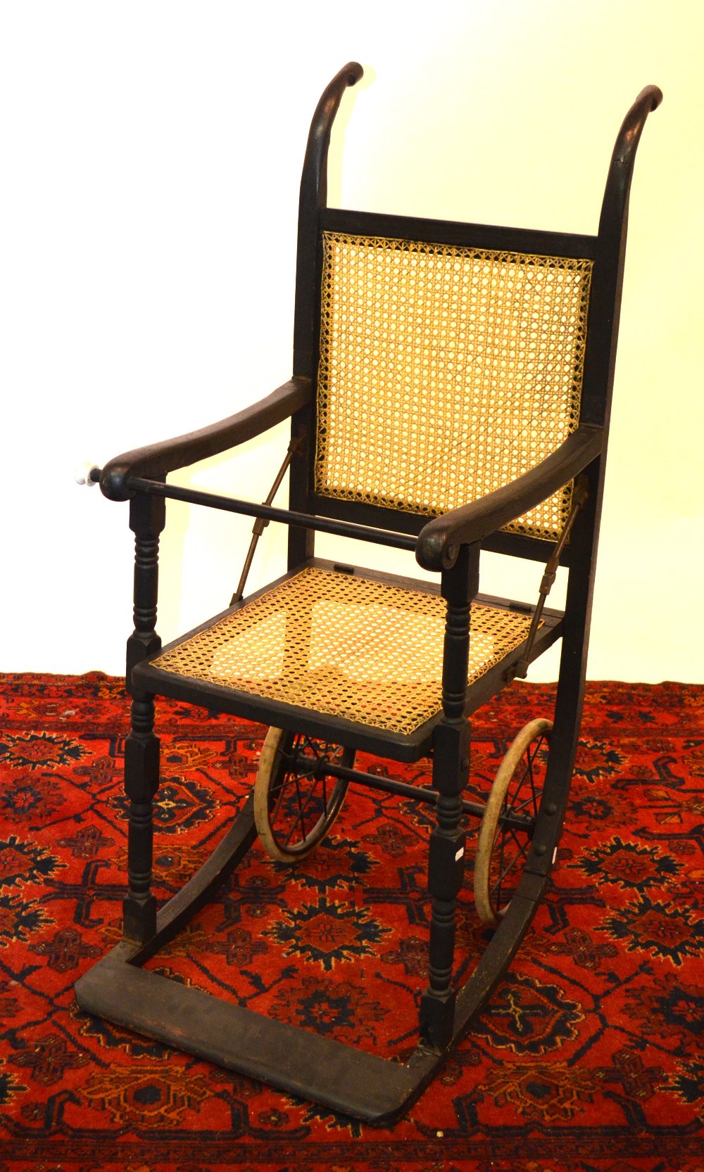An antique collapsable cane panelled ebonised invalid chair on spoked wheels