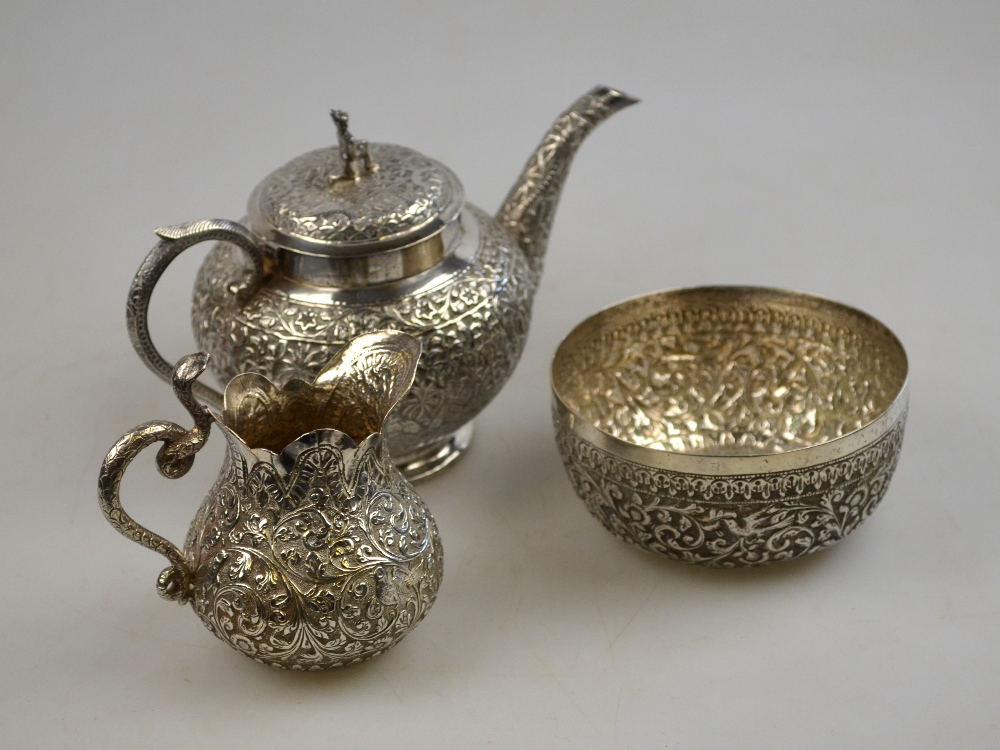 An Indian low grade white metal teapot, milk jug and sugar basin, each richly embossed and chased
