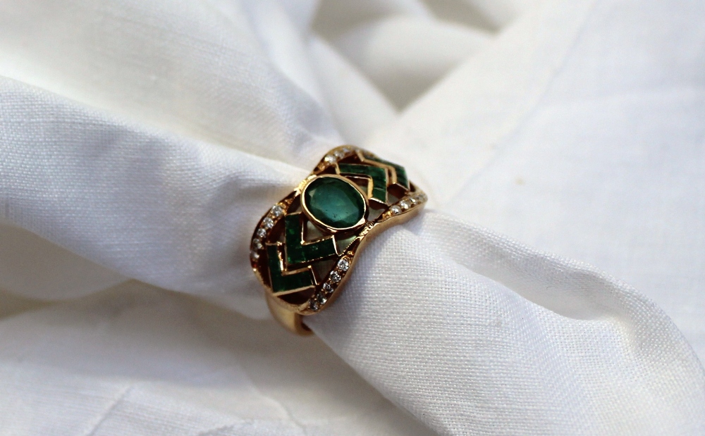 An emerald and diamond dress ring set with a central oval emerald flanked by princess cut emeralds