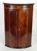 EARLY NINETEENTH CENTURY FIGURED MAHOGANY AND INLAID BOW FRONT CORNER CUPBOARD, the moulded cornice