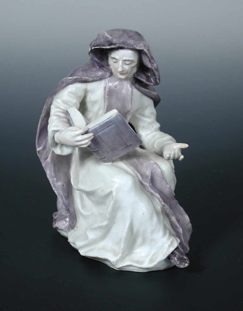 A Bow figure of an Abbess, she sits reading a book, her hooded cloak painted purple, 15.5cm (6 in)