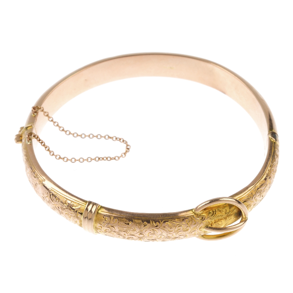 An Edwardian 9ct gold hinged bangle. The foliate engraved front panel, with stylised buckle, to the