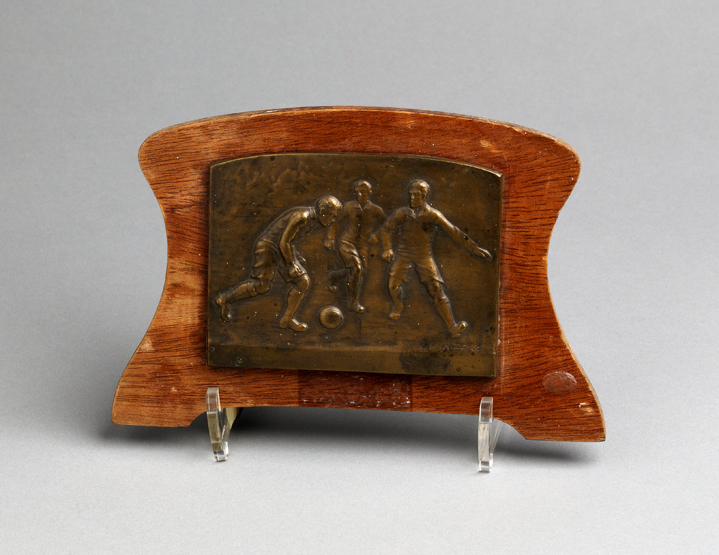 A bronze football plaque,
with a football scene signed H. Demey, mounted on a wooden backboard,