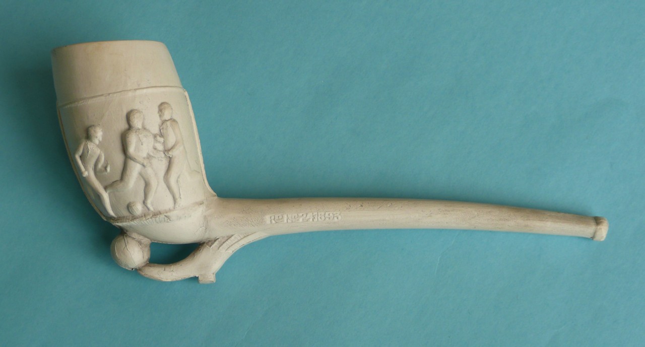 A clay pipe of large size moulded with a football scene, circa 1880, 227mm long, chip to foot