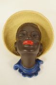 A Goldscheider pottery wall mask in the form of a black lady wearing a yellow sun hat and earring,
