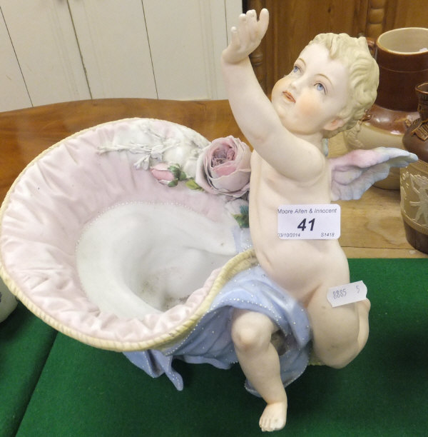 A 20th Century biscuit fired porcelain figure of a cherub beside a floral decorated straw hat