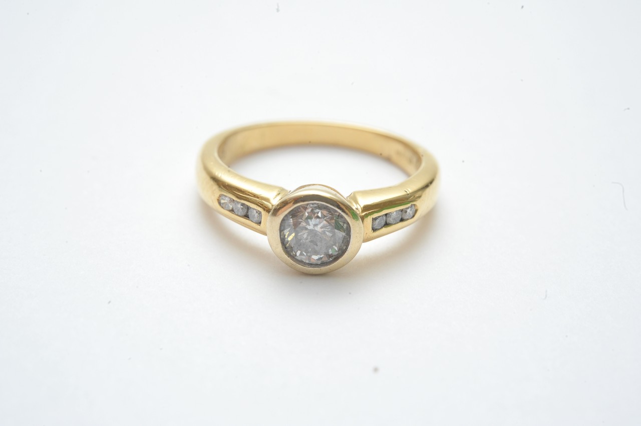 An 18ct yellow gold ring bezel set with a brilliant cut diamond, the shoulders each channel set with